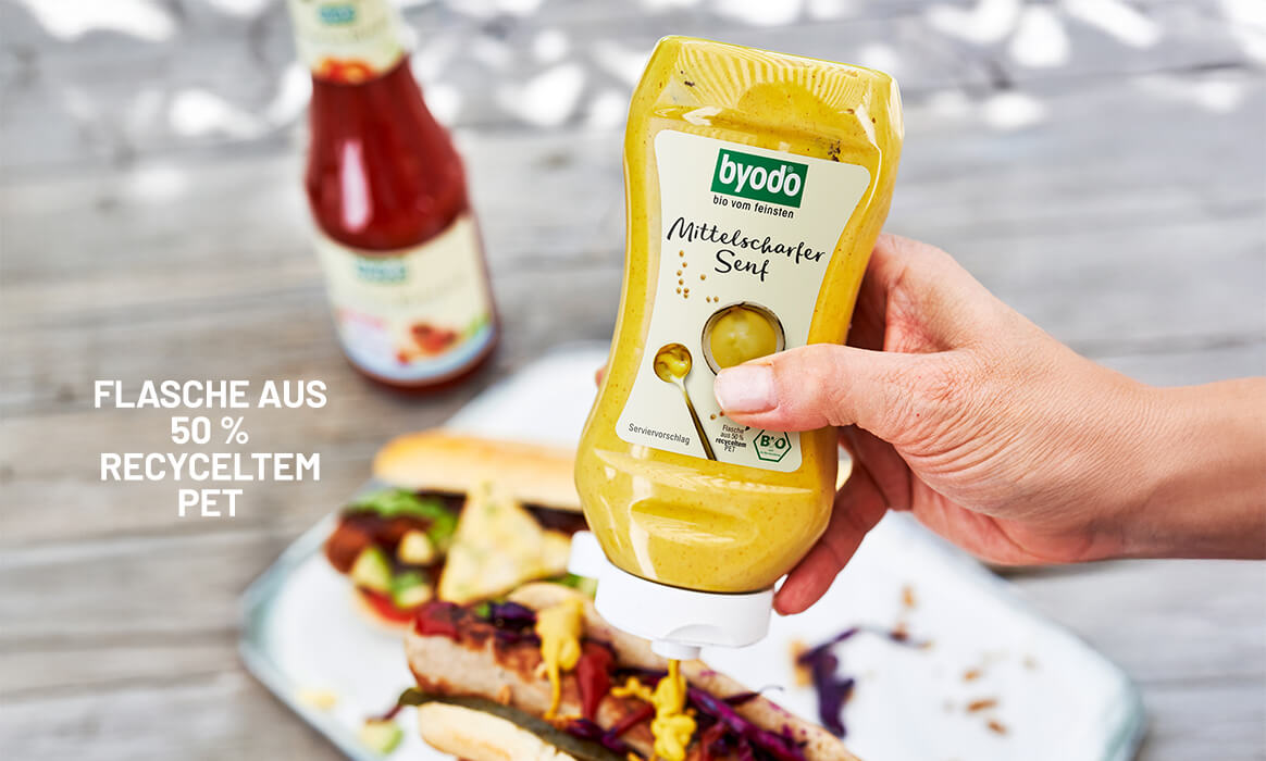 Byodo Mittelscharfer Senf in der PET Flasche im Hintergrund ein Hotdog in stimmungsvoller Umgebung