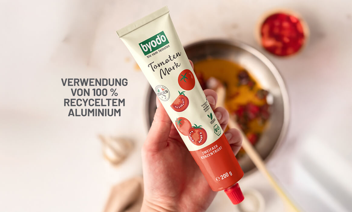Eine Hand hält das Byodo Tomatenmark in der Hand unscharf im Hintergrund eine Bratpfanne mit Zutaten